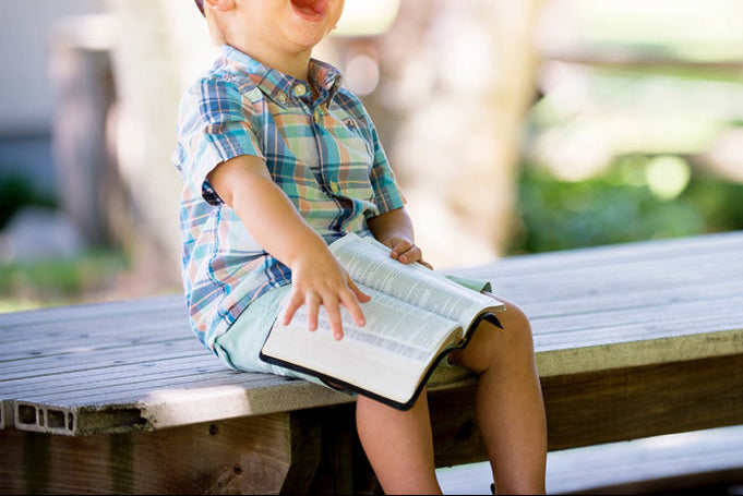 Child sitting on a wooden bench outdoors, holding a book and laughing.
